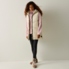 Persona con capelli ricci in piedi davanti a una parete chiara, indossa un gilet imbottito color crema, un maglione rosa, leggings neri e sneakers marroni, guarda verso il basso