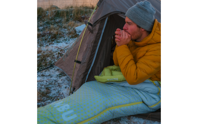 Person in yellow puffer jacket and gray beanie sits at a tent entrance with hands near the mouth; sleeping bag and lightly snow-covered ground visible