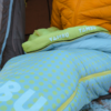 Close-up of a light green-blue sleeping bag pillow with polka dots and 'TAMBU' text, yellow jacket and dark tent fabric in the background, zipper and snow at the edge visible