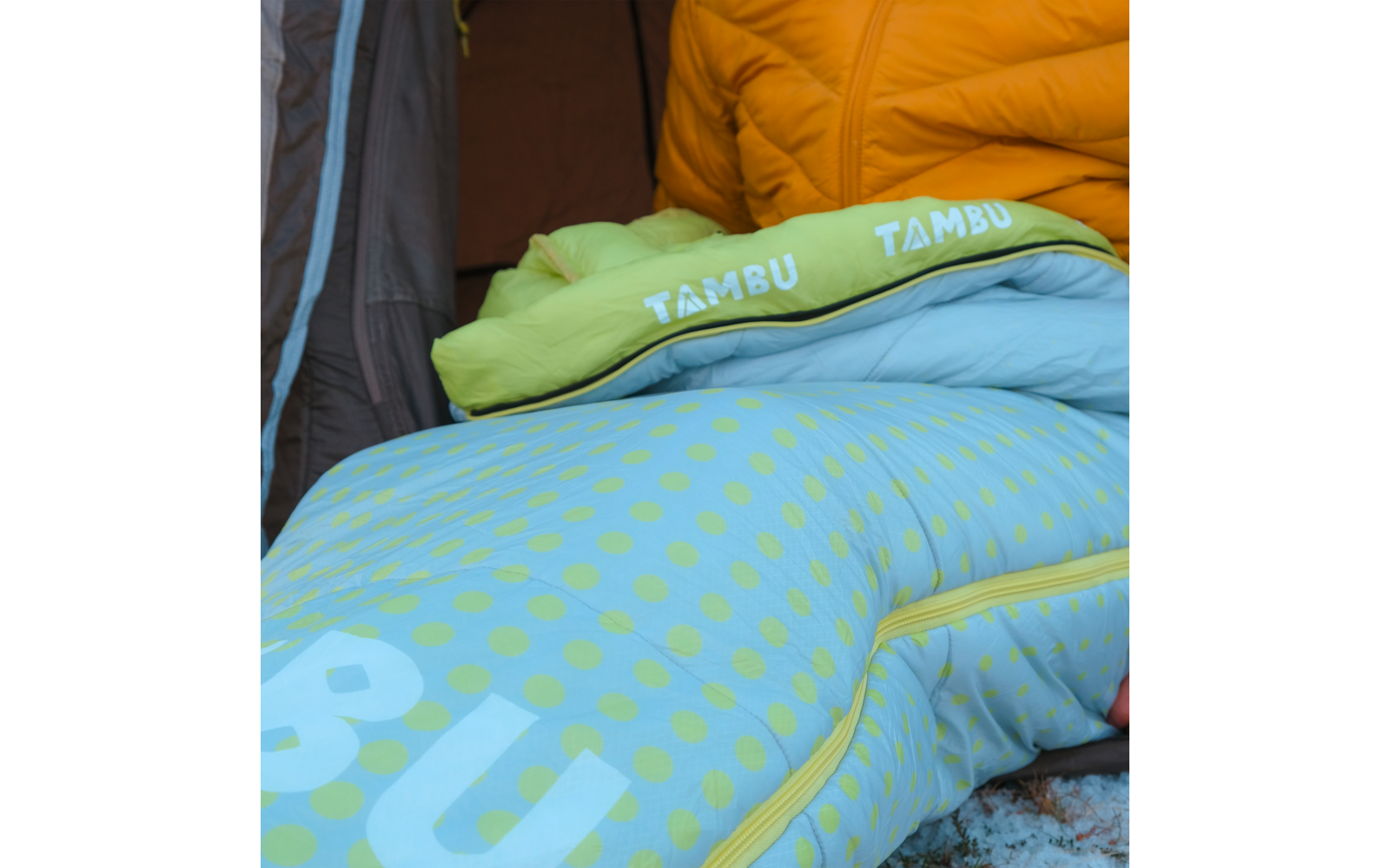 Close-up of a light green-blue sleeping bag pillow with polka dots and 'TAMBU' text, yellow jacket and dark tent fabric in the background, zipper and snow at the edge visible