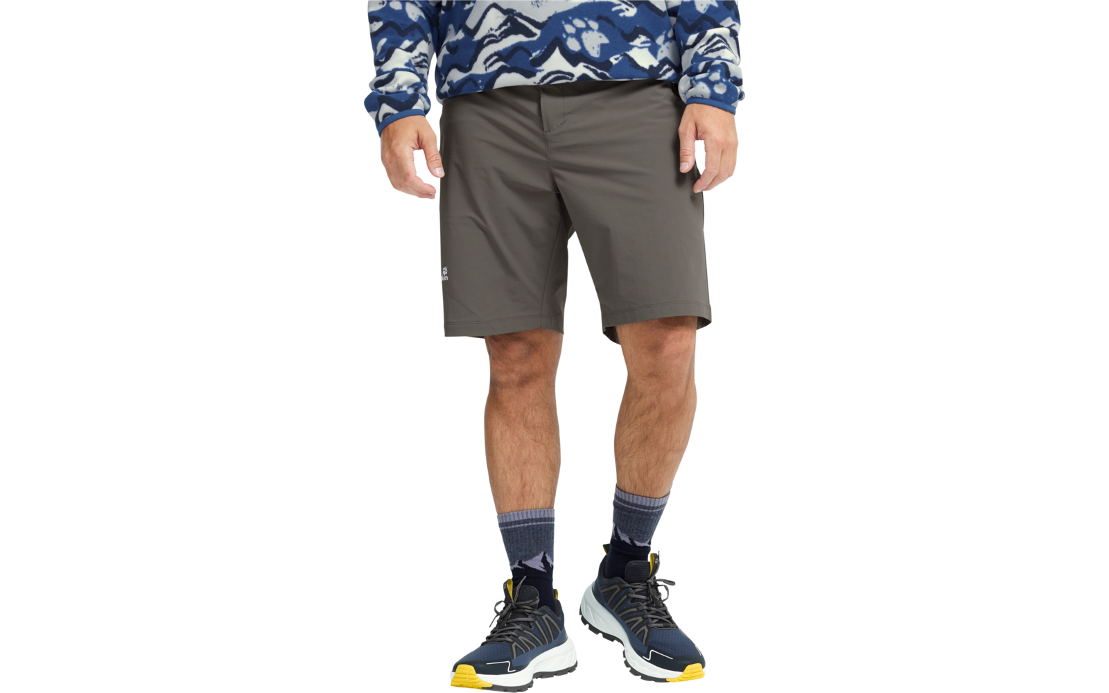 Persoon van taille tot voeten, draagt grijze knielange shorts, blauw-wit gemusterd trui tot taille zichtbaar, grijze sokken, blauwe sportschoenen met gele zolen; handen zijdelings zichtbaar