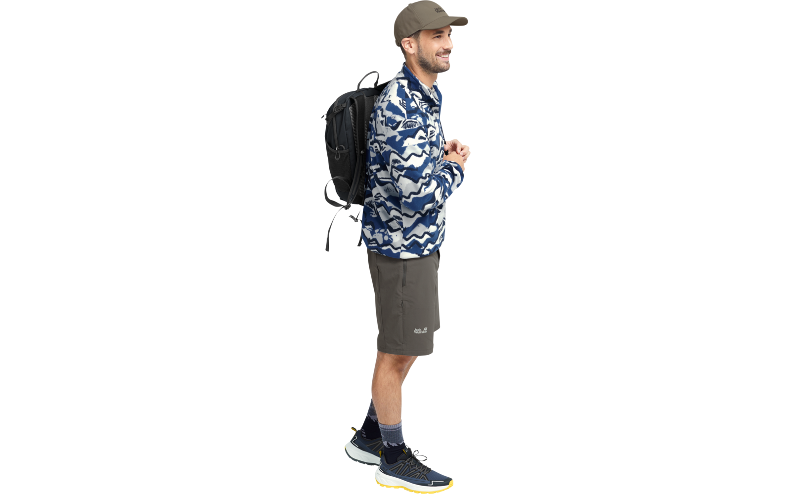 Man in zijaanzicht met baseballpet en korte baard, blauw-wit gemêleerde jas, grijze short, zwarte rugzak en sportschoenen