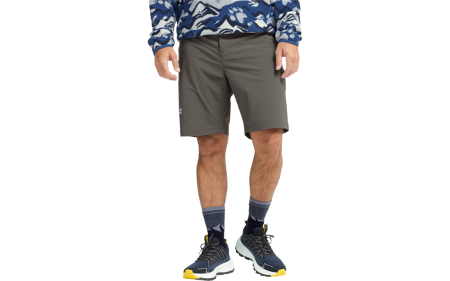 Persoon van taille tot voeten, draagt grijze knielange shorts, blauw-wit gemusterd trui tot taille zichtbaar, grijze sokken, blauwe sportschoenen met gele zolen; handen zijdelings zichtbaar