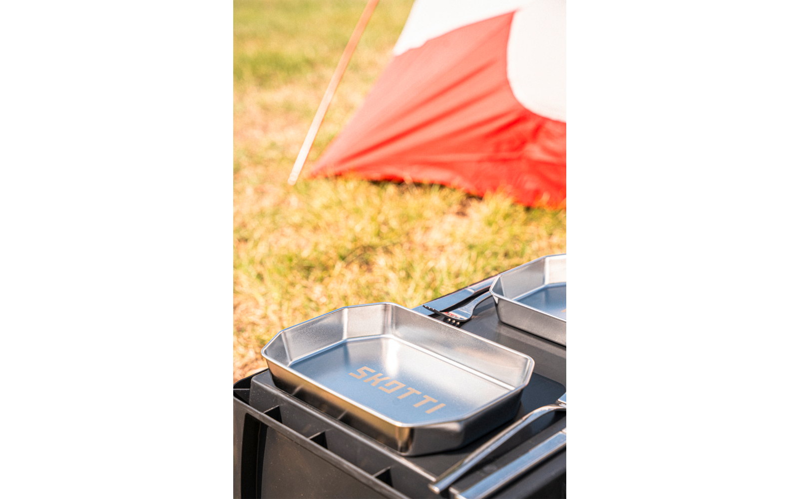 Metal pan with 'SKOTTI' lettering on a black tray, grass and a red-and-white tent in the background