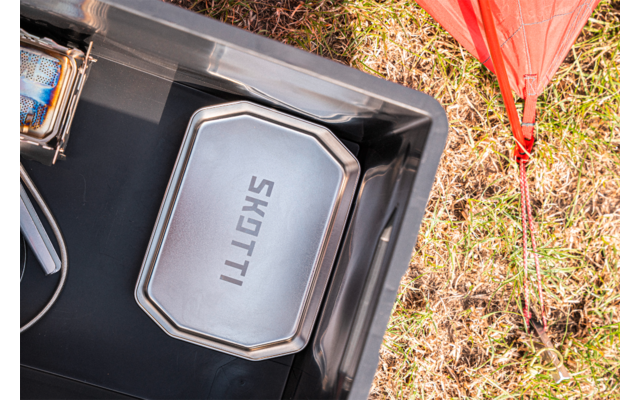 Metal box with the word "SKOTTI" on the lid inside a black plastic crate; to the right a red fabric triangle and grass
