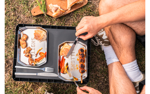 Two metal plates on a black tray with pieces of bread, carrot pieces, red pepper pieces and brown round balls; a hand holding a knife and fork, visible legs wearing sneakers and white socks; wooden board with bread and herbs on grass