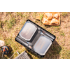Two silver rectangular food containers in a black plastic crate, two forks between the lids, metal grill grate, small gas canister with hose, wooden tray with four bread rolls, grassy ground, part of a shoe visible