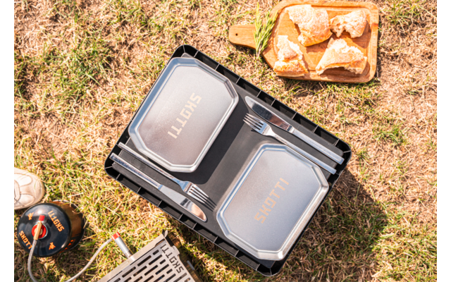 Two silver rectangular food containers in a black plastic crate, two forks between the lids, metal grill grate, small gas canister with hose, wooden tray with four bread rolls, grassy ground, part of a shoe visible