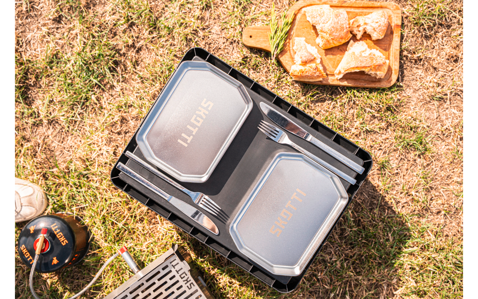 Two silver rectangular food containers in a black plastic crate, two forks between the lids, metal grill grate, small gas canister with hose, wooden tray with four bread rolls, grassy ground, part of a shoe visible
