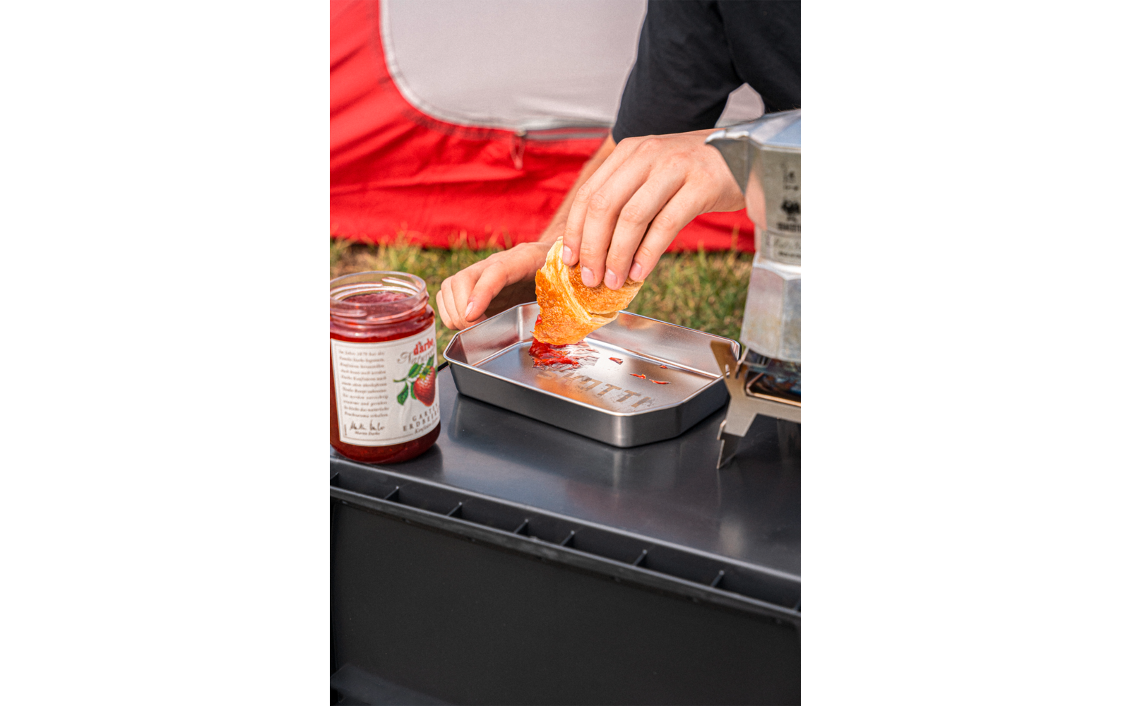 Hand holding a triangular tortilla chip dipping it into a small metal container with orange-colored spread; a jar of red jam beside it; a red tent on grass in the background