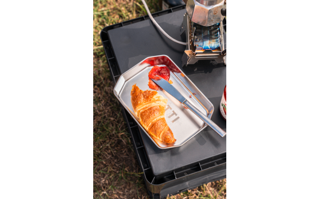 Croissant on a metal tray, knife with red jam, jar of jam at the edge, metal appliance and black plastic crate on grass