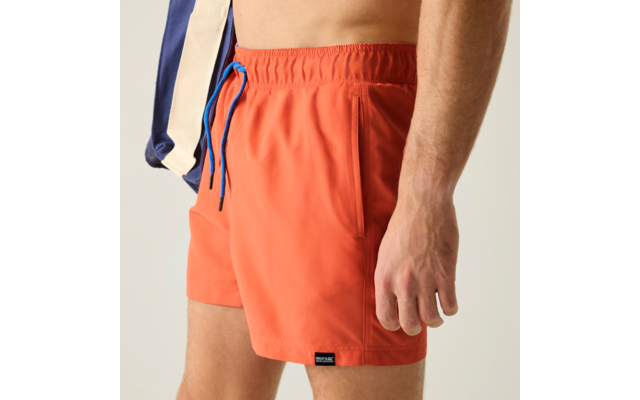 Unterer Torso und Oberschenkel einer Person in orangefarbenen Badeshorts mit blauem Kordelzug, rechte Hand seitlich, sichtbare Seitentasche, Teil einer dunkelblauen und beigen Jacke über der Schulter