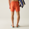 Rückenansicht einer Person von der Taille abwärts, orange Badeshorts, blau-weiß gestreifte Tasche über der Schulter, weiße Sandalen, heller einfarbiger Hintergrund