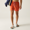 Unterkörper einer Person mit oranger Badeshorts und blauem Kordelzug, hält blau-beige gestreiften Stoff, weiße Sandalen mit blauen Sohlen, heller Hintergrund