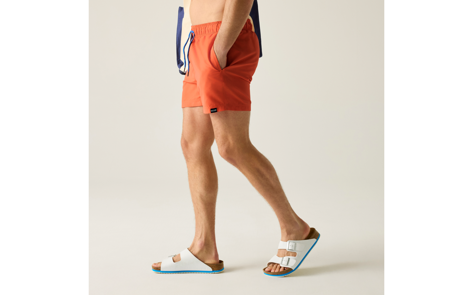 Seitliche Ansicht der Beine und Hüften einer Person in orangefarbenen Badeshorts, Hände in den Taschen, weiße Sandalen mit zwei Riemen und blauer Sohle, blauer Stoffstreifen an der Hüfte, heller Hintergrund