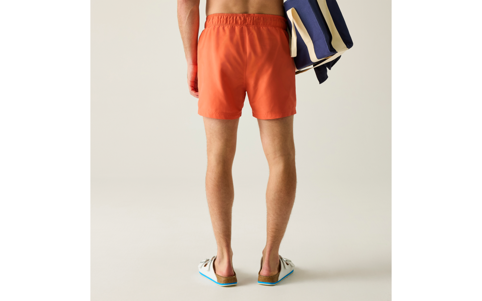 Rückenansicht einer Person von der Taille abwärts, orange Badeshorts, blau-weiß gestreifte Tasche über der Schulter, weiße Sandalen, heller einfarbiger Hintergrund