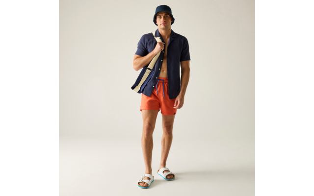 Person steht vor hellem neutralem Hintergrund, trägt dunkelblauen Fischerhut, offenes dunkelblaues Kurzarmhemd, orangefarbene kurze Hose, weiße Sandalen und eine dunkelblaue Tragetasche mit hellen Streifen über der Schulter
