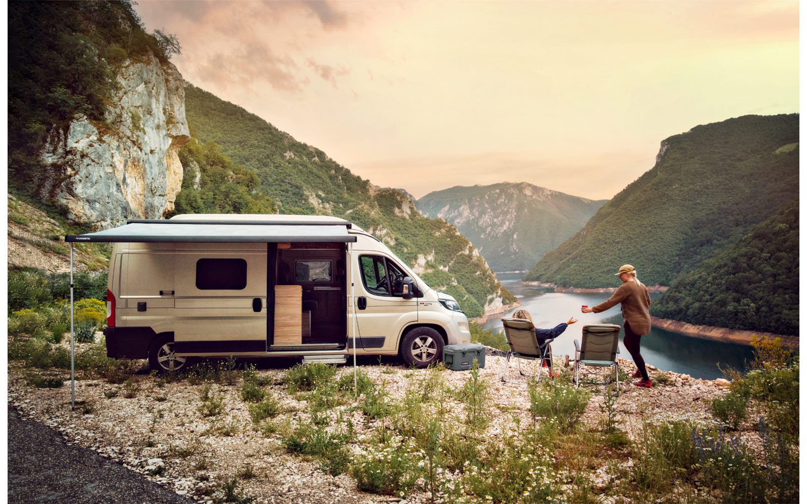 Camper van parked by a riverbank with mountains in the background, man casting fishing line
