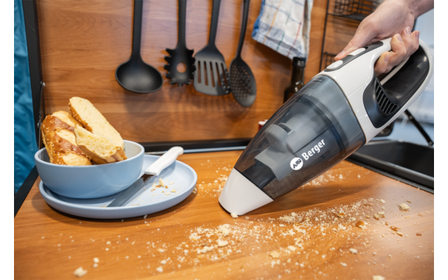 Main tenant un aspirateur portable au-dessus de miettes de pain sur un plan de travail en bois ; à côté un bol bleu avec des tranches de pain sur une assiette, des ustensiles de cuisine accrochés au mur et un torchon