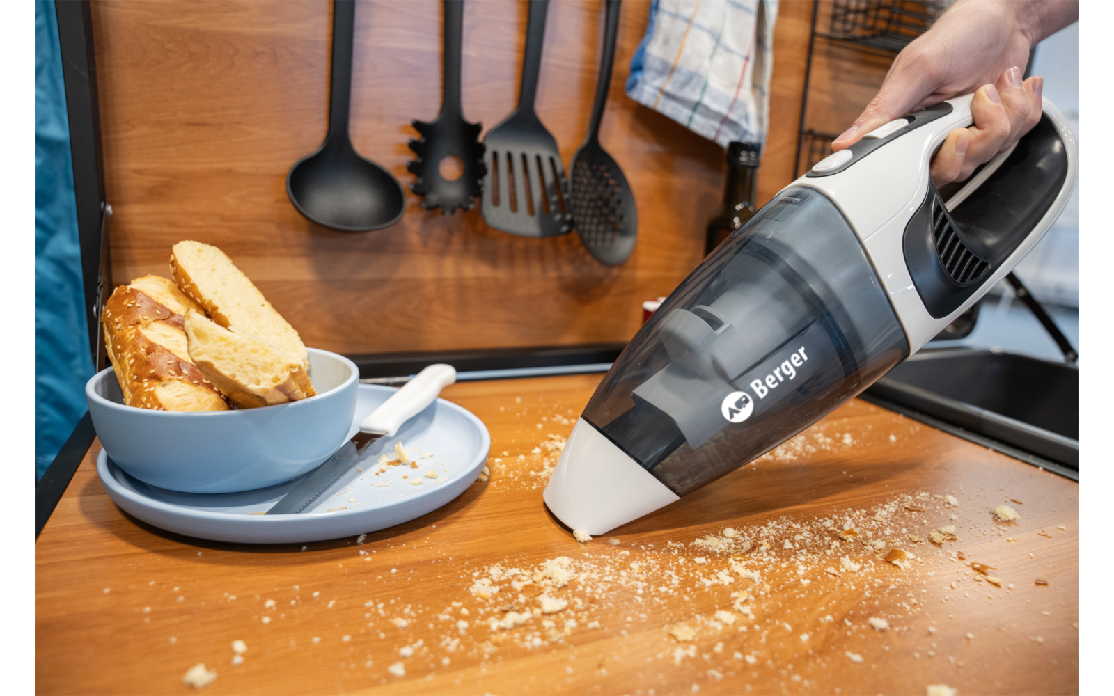 Main tenant un aspirateur portable au-dessus de miettes de pain sur un plan de travail en bois ; à côté un bol bleu avec des tranches de pain sur une assiette, des ustensiles de cuisine accrochés au mur et un torchon