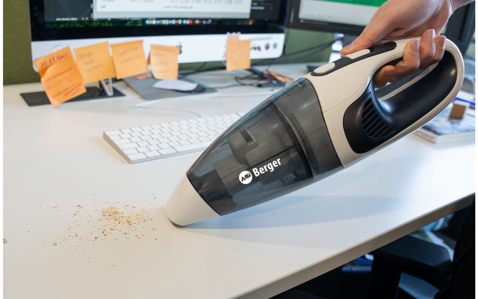 Main tenant un aspirateur à main au‑dessus de miettes sur un bureau blanc ; clavier, deux écrans et des post‑it en arrière‑plan