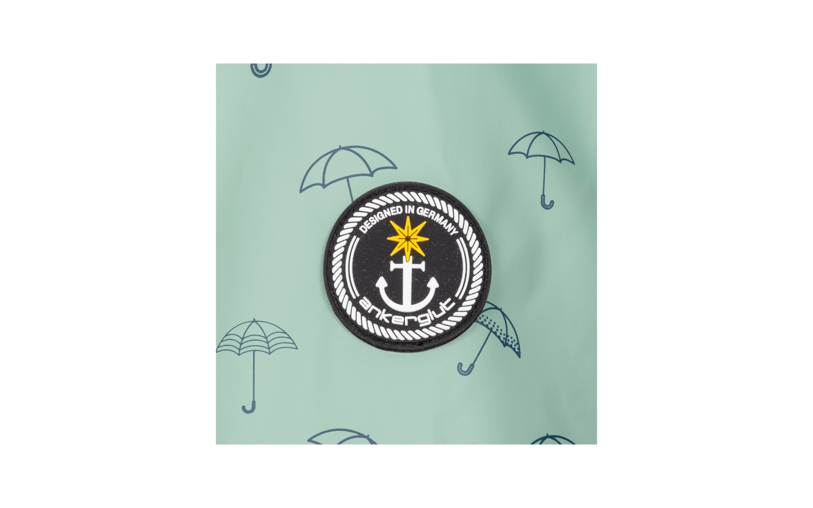 Ronde zwarte patch met witte anker, gele ster erboven, witte touwrand en tekst 'DESIGNED IN GERMANY' en 'ankerglück' op een mintgroene achtergrond met kleine paraplu-illustraties