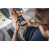 Personne assise sur une chaise longue tenant un smartphone affichant un écran d'application bleu avec un cadran circulaire, porte un t‑shirt bleu foncé et un short beige ; tablette noire sur l'accoudoir visible