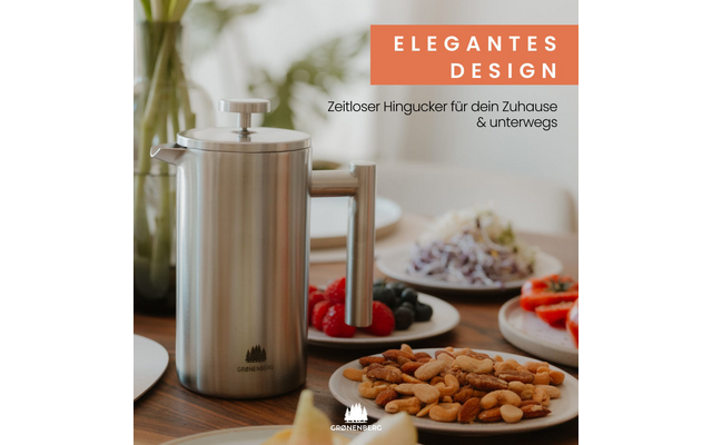 Prensa francesa de acero inoxidable sobre mesa de madera, cuenco de frutos secos en primer plano, platos con bayas, ensalada y rodajas de tomate al fondo, botella de vidrio y planta a la izquierda, arriba a la derecha texto ELEGANTES DESIGN y Zeitloser Hingucker für dein Zuhause & unterwegs, logotipo de la marca abajo