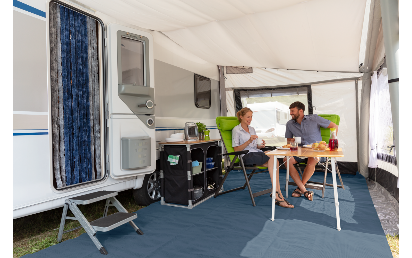 Zwei Personen sitzen in der Vorzeltfläche neben einem geparkten Wohnwagen an einem kleinen Tisch; Teller, Getränke und Obst auf dem Tisch; grüne Klappstühle, mobile Campingküche und geöffnete Wohnwagentür sichtbar; blauer Bodenbelag unter den Möbeln