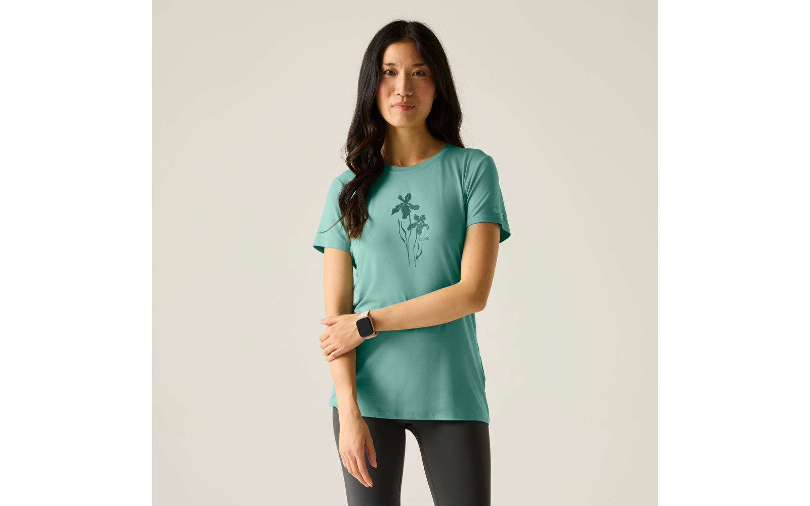 Frau mit dunklen Haaren, grünes T-Shirt mit Blumenmotiv, schwarze Leggings, steht vor hellem Hintergrund, schaut in die Kamera, Arme vor dem Körper