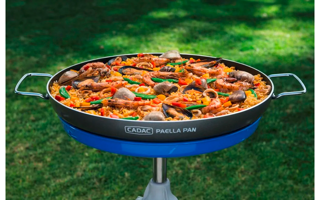 Paellapan op standaard op groen gras, gevuld met gele rijst, garnalen, mosselen, rode paprika-repen en lichtbruine vleesstukjes