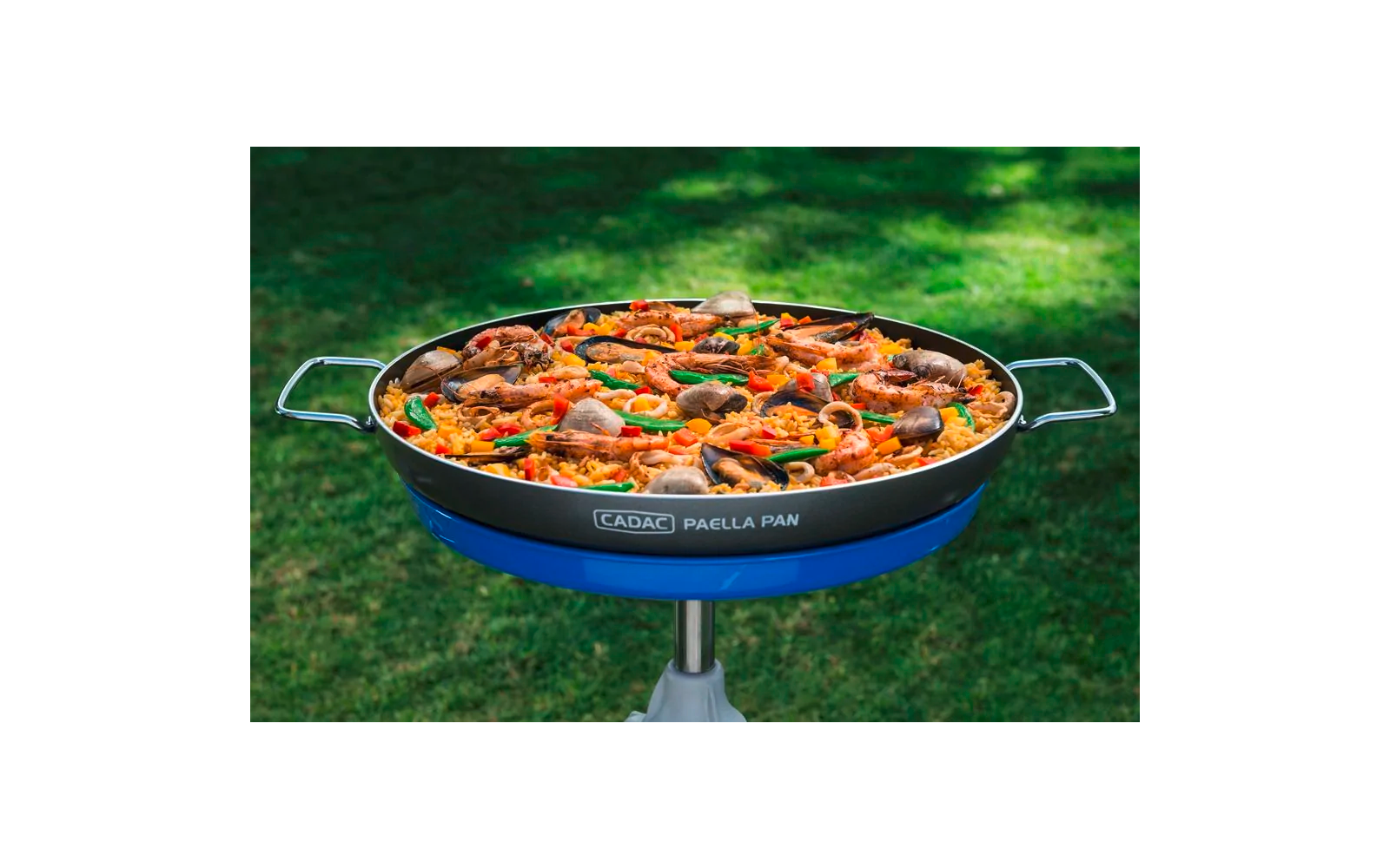Paellapan op standaard op groen gras, gevuld met gele rijst, garnalen, mosselen, rode paprika-repen en lichtbruine vleesstukjes