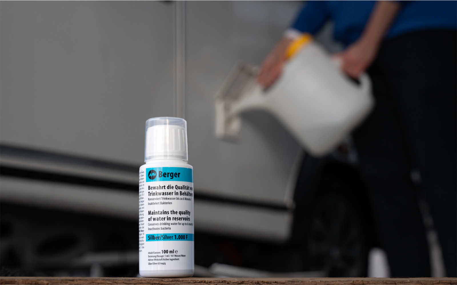 Bouteille blanche avec étiquette sur une surface en bois au premier plan ; à l'arrière-plan une personne floue en tenue bleue avec un gant jaune verse du liquide d'un récipient blanc dans une ouverture d'un véhicule