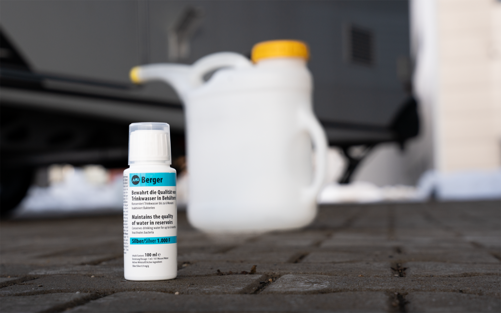 Petite bouteille blanche avec étiquette sur sol pavé, en arrière-plan flou grand bidon blanc en plastique avec bouchon jaune, partie d'un véhicule et mur de bâtiment visibles, traces de neige