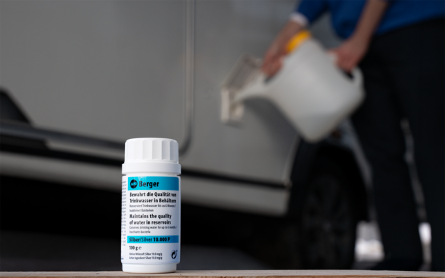 Bouteille blanche avec étiquette bleue au premier plan ; personne floue verse depuis un récipient blanc dans l'orifice de remplissage d'une voiture argentée