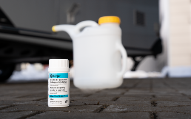 Petite bouteille blanche avec étiquette sur un sol pavé; en arrière-plan un bidon plastique blanc plus grand à bouchon jaune, à gauche un pneu de voiture, traces de neige