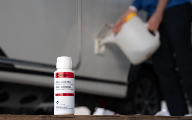 Petite bouteille blanche avec étiquette rouge au premier plan sur une surface en bois, à l'arrière-plan une personne floue en haut bleu penche un bidon blanc et verse du liquide à côté d'une voiture garée gris clair