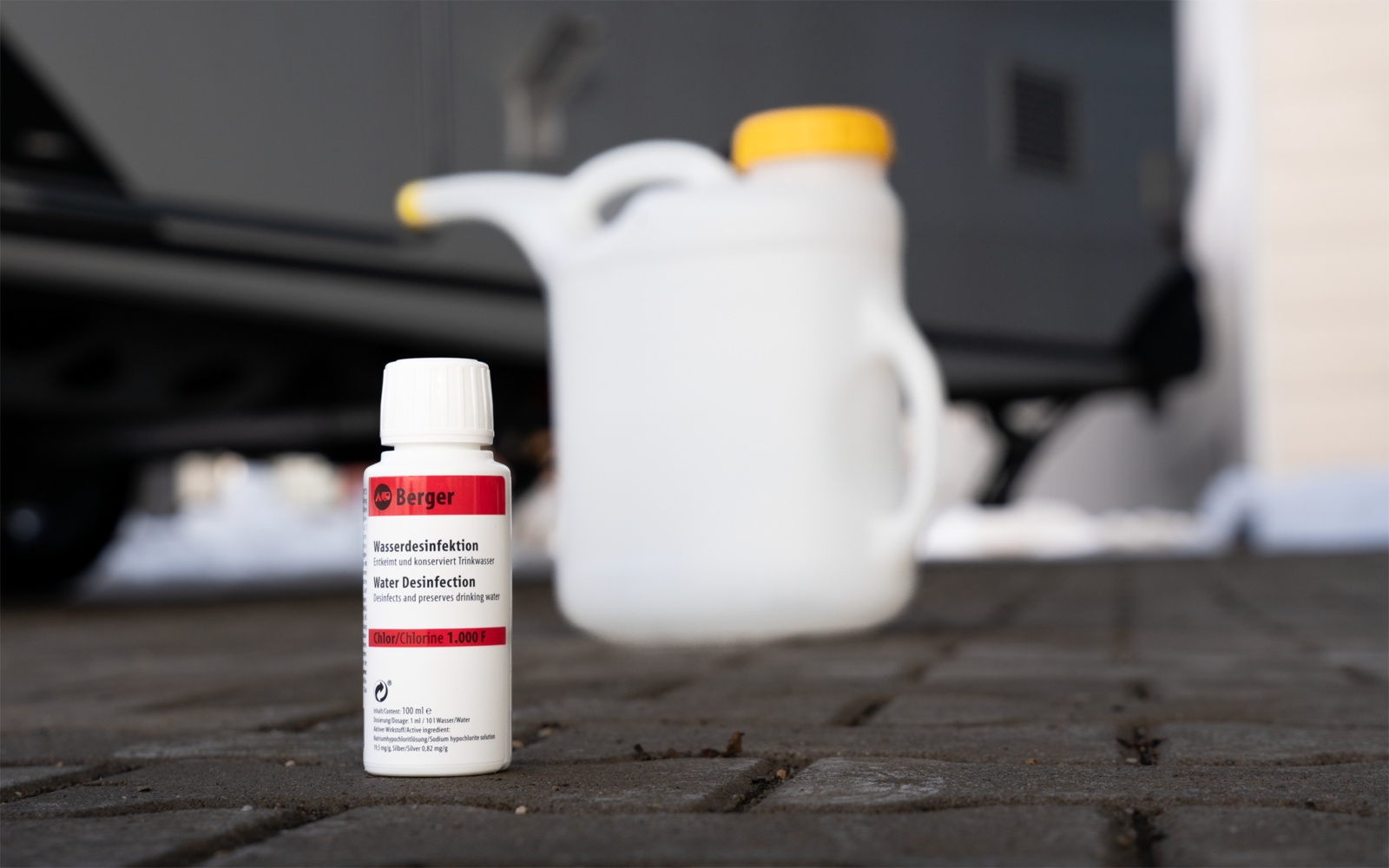 Petite bouteille blanche avec étiquette rouge et bouchon blanc sur pavé ; en arrière-plan un bidon plastique blanc flou avec bouchon jaune, une roue de véhicule et de la neige