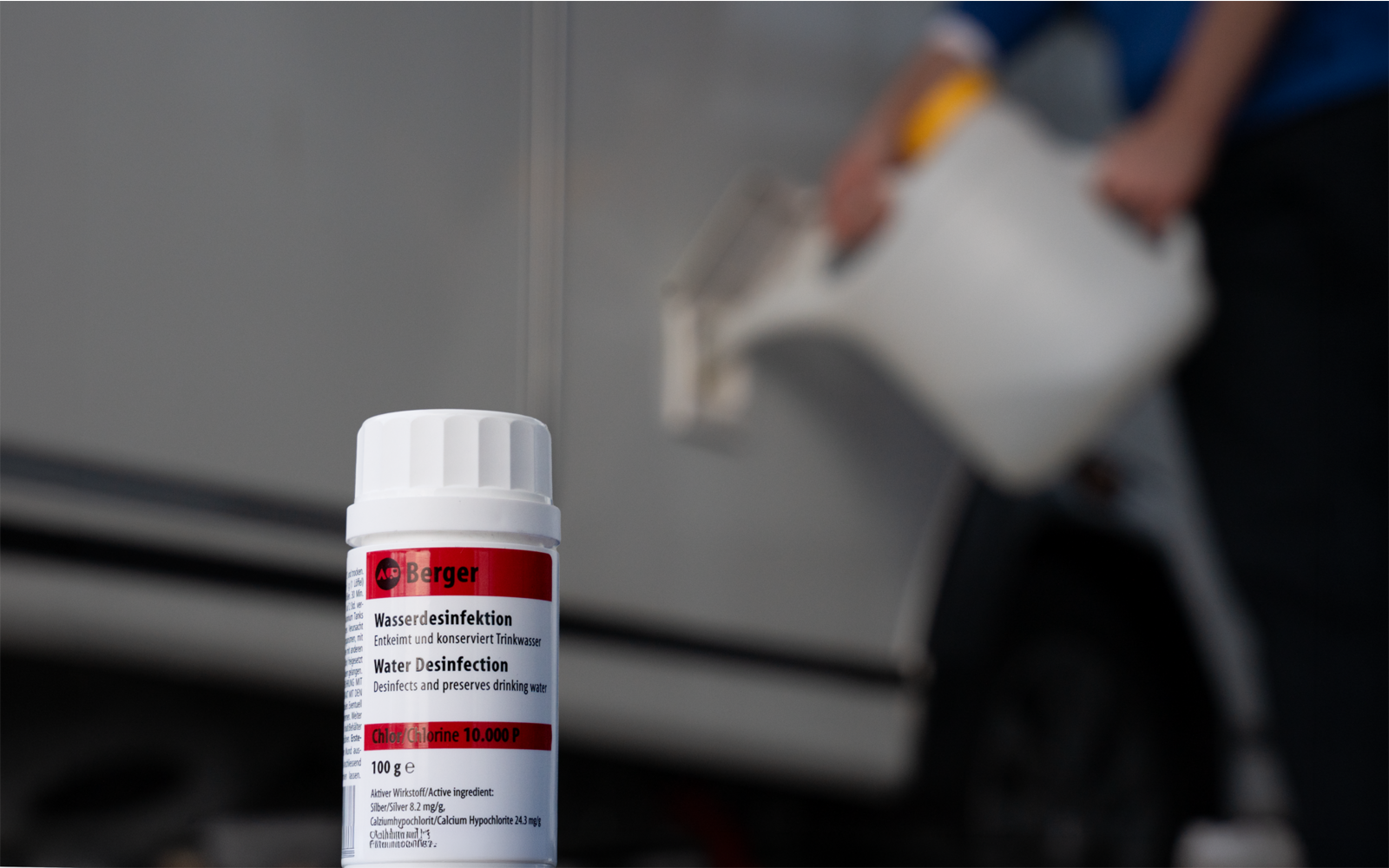 Bouteille en plastique blanche avec bouchon à vis et étiquette rouge au premier plan ; en arrière-plan flou, une personne en haut bleu verse d'un arrosoir blanc dans une ouverture sur le côté d'un véhicule gris