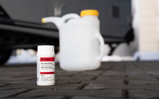 Petit flacon plastique blanc avec étiquette rouge sur sol pavé ; à l'arrière-plan flou un bidon blanc avec bouchon jaune, un véhicule au coffre ouvert et un mur de bâtiment