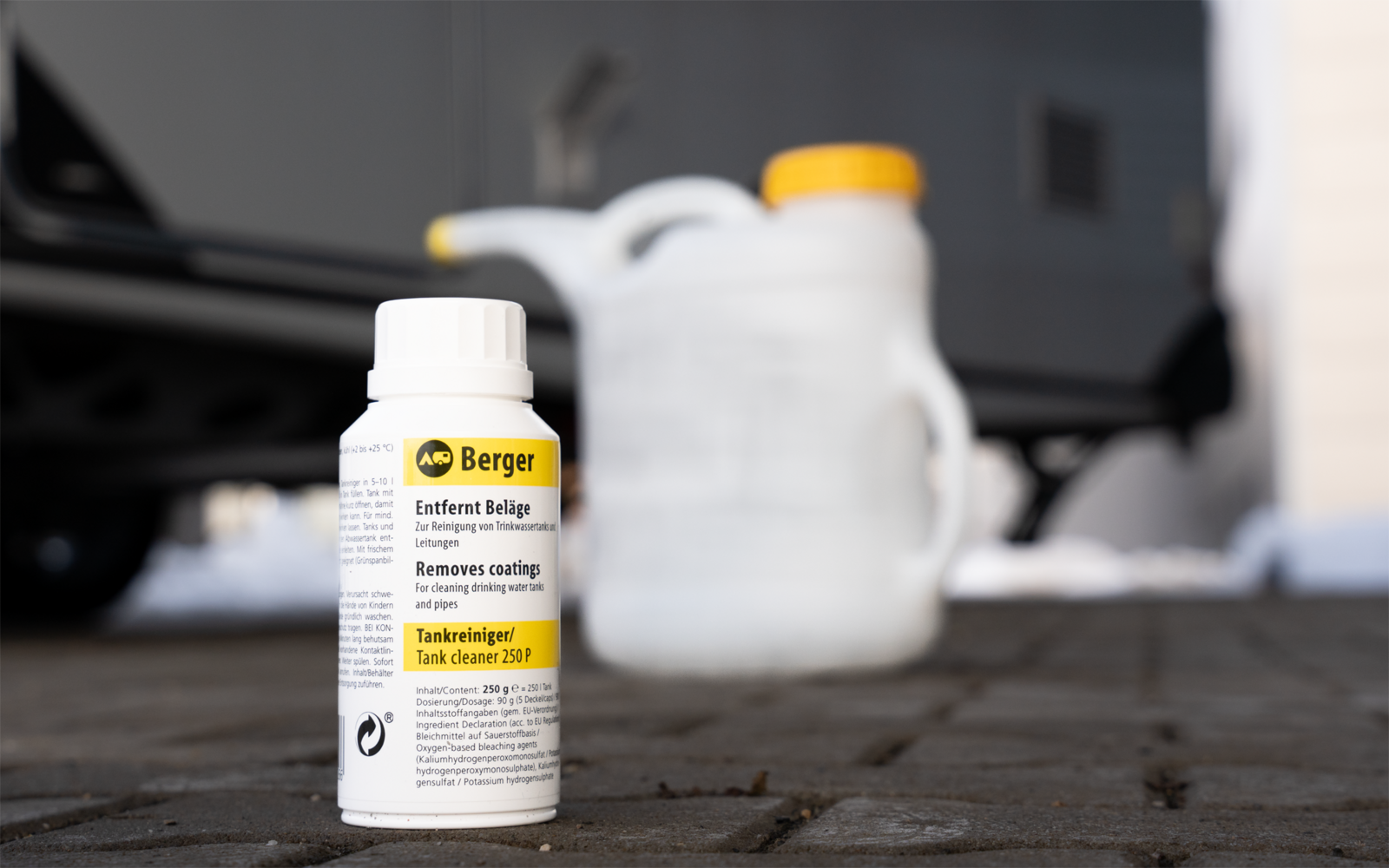 Kleine weiße Flasche mit weißem Verschluss und gelb-schwarzem Etikett mit der Aufschrift 'Berger' auf gepflastertem Boden; im Hintergrund zwei weiße Kanister mit gelben Verschlüssen und ein unscharfes Fahrzeug
