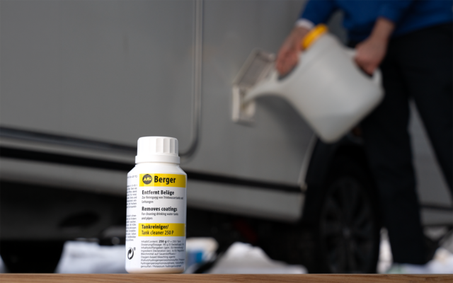 Weiße Kunststoffflasche mit gelbem Etikett auf einer Holzfläche im Vordergrund; im Hintergrund unscharfe Person gießt Flüssigkeit aus einem weißen Kanister an der Seite eines silbernen Autos, schwarzer Reifen sichtbar