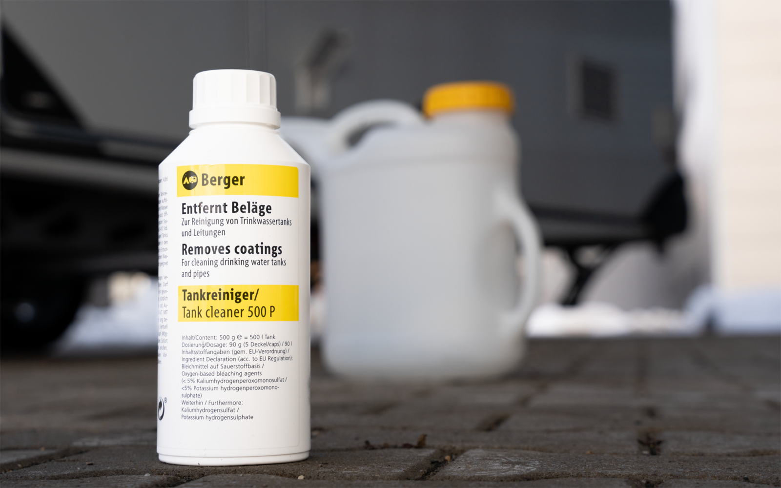 Weiße Plastikflasche mit gelbem Etikett ('Berger Entfernt Beläge') im Vordergrund; unscharfe weiße Kanister mit gelbem Verschluss, Papierrolle rechts; gepflasterter Boden und unscharfes Auto im Hintergrund