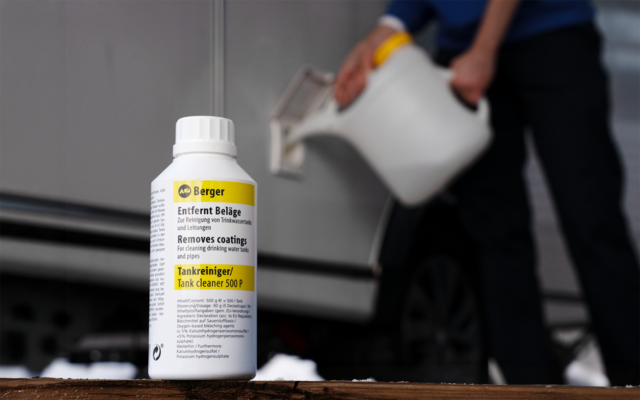 Weiße Kunststoffflasche mit Etikett auf einer Holzoberfläche im Vordergrund; im Hintergrund unscharfe Person gießt Flüssigkeit aus einem weißen Kanister neben einem grauen Auto; Schneereste auf dem Boden