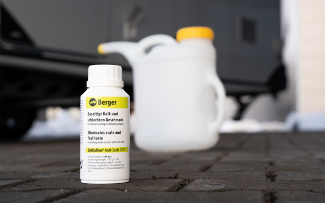 Weiße Kunststoffflasche mit 'Berger'-Etikett auf gepflastertem Boden; im Hintergrund unscharfer weißer Kanister mit gelbem Verschluss, verschwommene Fahrzeugteile und Schneereste