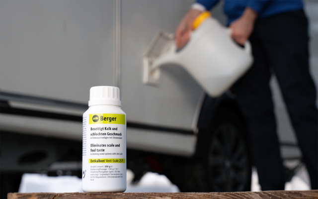 Weiße Flasche mit gelb‑schwarzem Etikett auf einer Holzfläche im Vordergrund; im Hintergrund unscharf gießt eine Person Flüssigkeit aus einem weißen Kanister in die geöffnete Tankklappe eines silbernen Autos