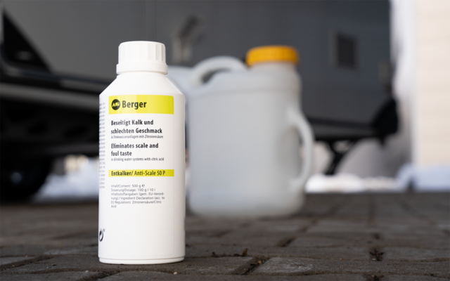 Weiße Kunststoffflasche mit Etikett 'Berger' auf gepflastertem Boden, verschwommene weiße Kanister und Teil eines Autoreifens im Hintergrund