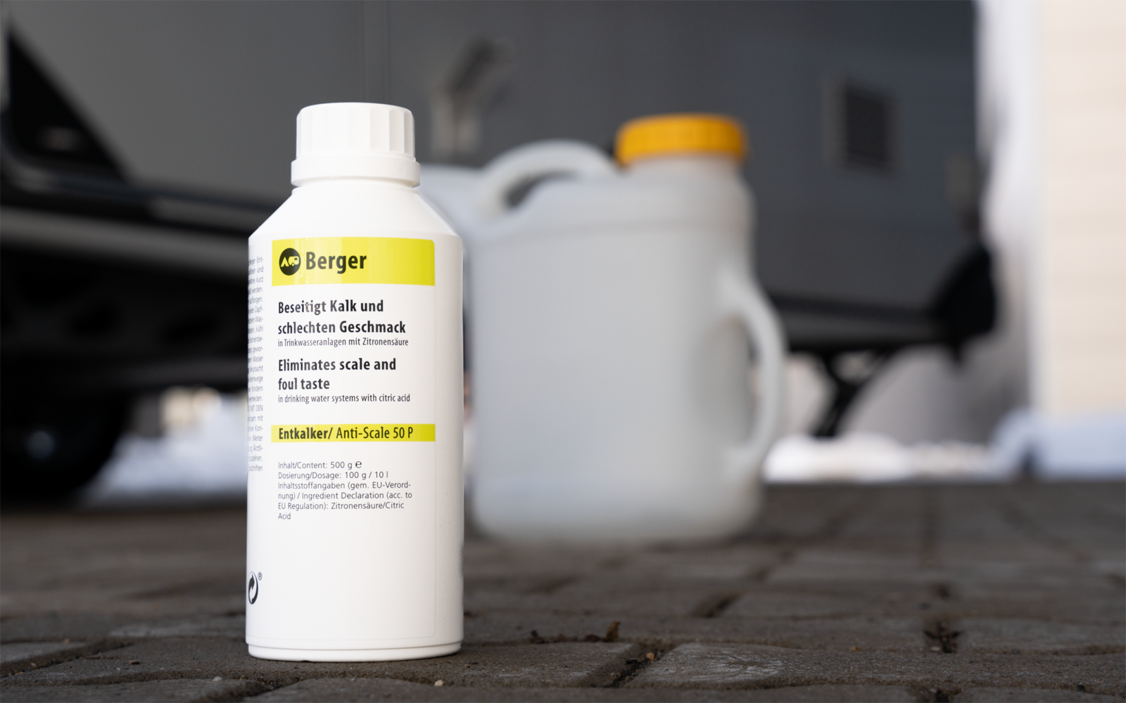 Weiße Kunststoffflasche mit Etikett 'Berger' auf gepflastertem Boden, verschwommene weiße Kanister und Teil eines Autoreifens im Hintergrund