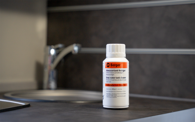 Weiße Kunststoffflasche mit orange-weißem Etikett 'Berger' auf Arbeitsplatte neben Spüle und verchromtem Wasserhahn, dunkle Fliesen im Hintergrund