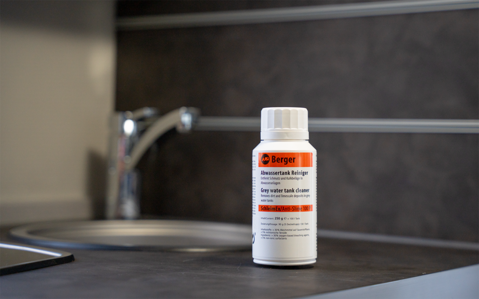 Weiße Kunststoffflasche mit orange-weißem Etikett 'Berger' auf Arbeitsplatte neben Spüle und verchromtem Wasserhahn, dunkle Fliesen im Hintergrund